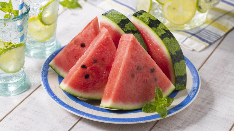 Several slices of watermelon on a plate, sitting on a table with several glasses of refreshing-looking beverages surrounding it