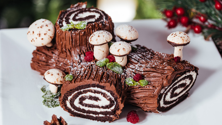 A festive, decorated Yule log cake on a white plate in front of a Christmas tree