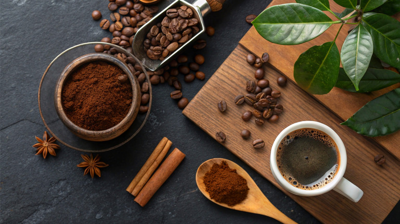 Coffee in a ceramic mug, loose whole spices, ground coffee in a bowl, loose coffee beans in a metal scoop on a table and wooden cutting board.