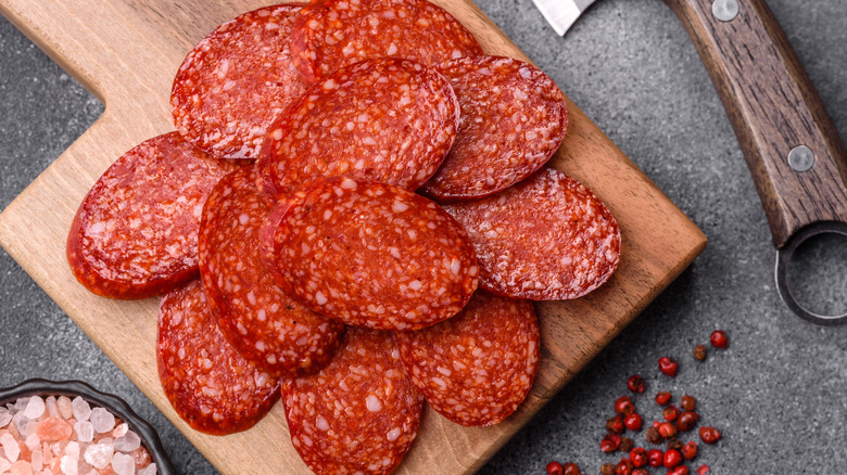 Slices of fresh pepperoni on a cutting board with peppercorns, rock salt, and a knife nearby