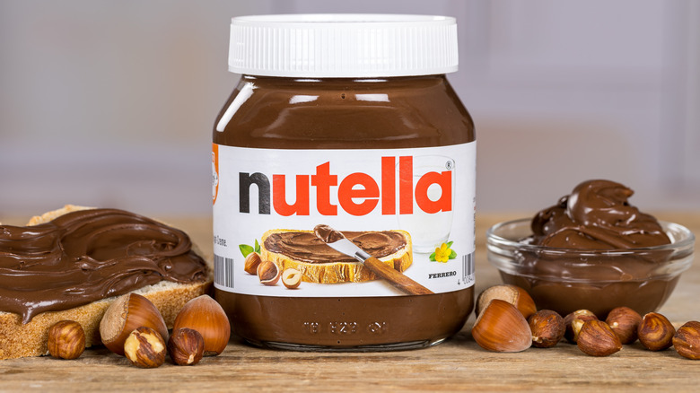 A large jar of Nutella in the center, flanked by hazelnuts and bread with the spread