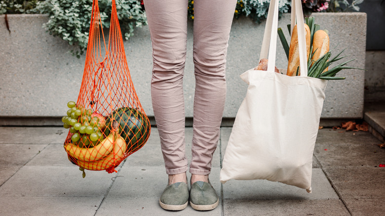 two reusable grocery bags loaded with produce and bread held beside a person's legs