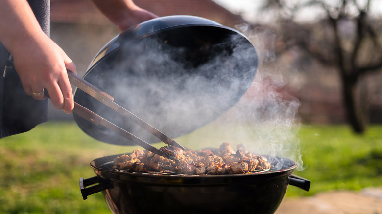 person lifting lid off kettle grill and moving meat with grill tongs
