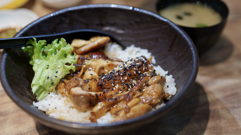 a bowl of teriyaki chicken garnished with sesame seeds over rice, with miso soup in the background