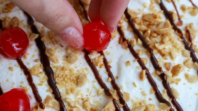 Someone placing a maraschino cherry on top of whipped cream covered in crushed peanuts and a chocolate drizzle