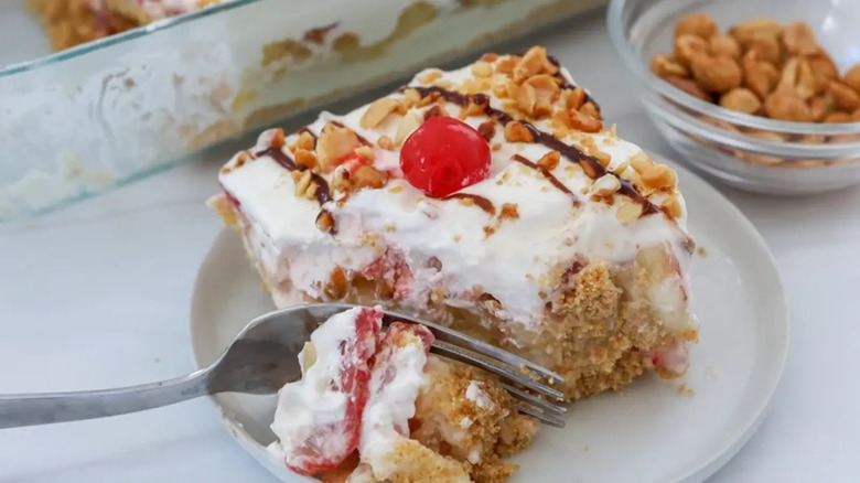 A fork cutting into a slice of no-bake banana split cake topped with whipped cream, chocolate drizzle, peanuts, and a maraschino cherry