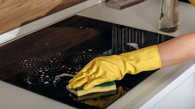 A person wearing a yellow rubber glove using a sponge to clean a black glass stove top
