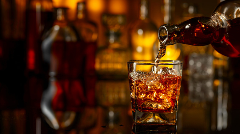 Bourbon being poured from a glass bottle into a glass tumbler on a bar-top, with other liquor bottles in the background