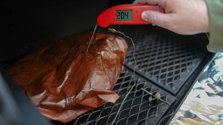 Someone places a meat thermometer into a coovered brisket on a smoker