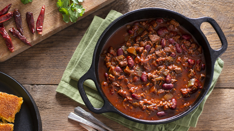 A skillet of chili with dried chillies on a board besides
