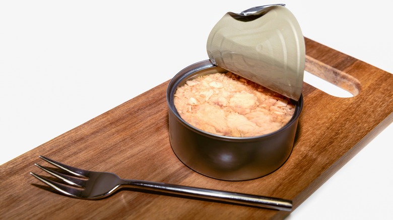 An open can of tuna on a wooden cutting board next to a fork