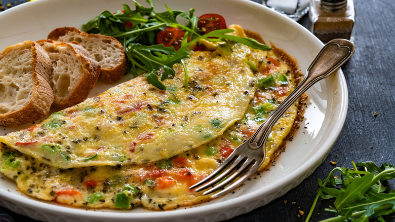 A vegetable omelet folded on a white plate, garnished with toasted bread and an arugula and tomato salad