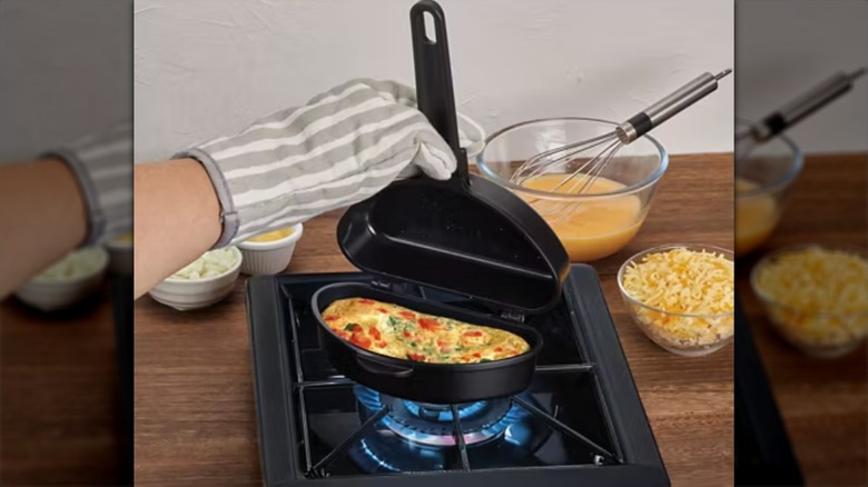 A person wearing a striped oven mitt using Aldi's Crofton omelet pan to make an omelet on their stovetop