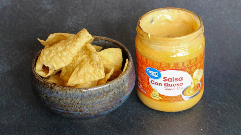 Open jar of Great Value Salsa Con Queso next to stoneware bowl of tortilla chips on dark surface