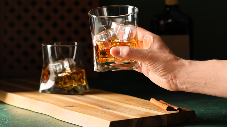 Person holding a glass of whiskey with ice cubes above a board.