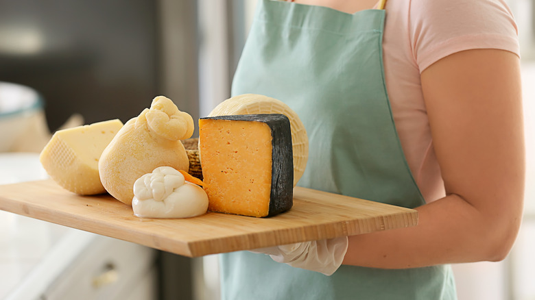 An aproned woman hols a wooden tray filled with different cheeses