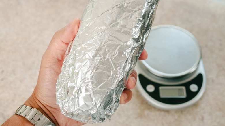 A person holds a foil wrapped burrito over a scale