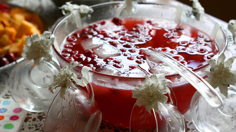 A vintage punch bowl full of cranberry punch and lined with matching glasses adorned with white flowers and a large clear plastic ladle resting in the bowl