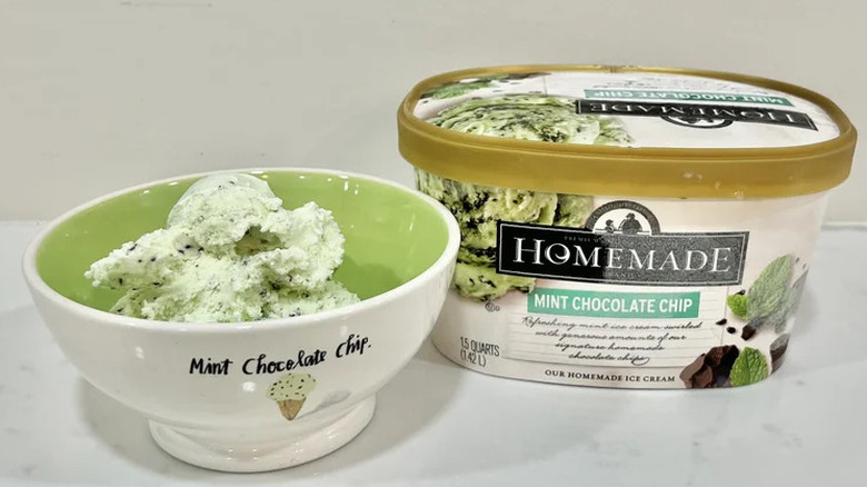 A tub of Homemade Brand Mint Chocolate Chip ice cream sitting on a white surface, before a white background, next of a bowl of the ice cream
