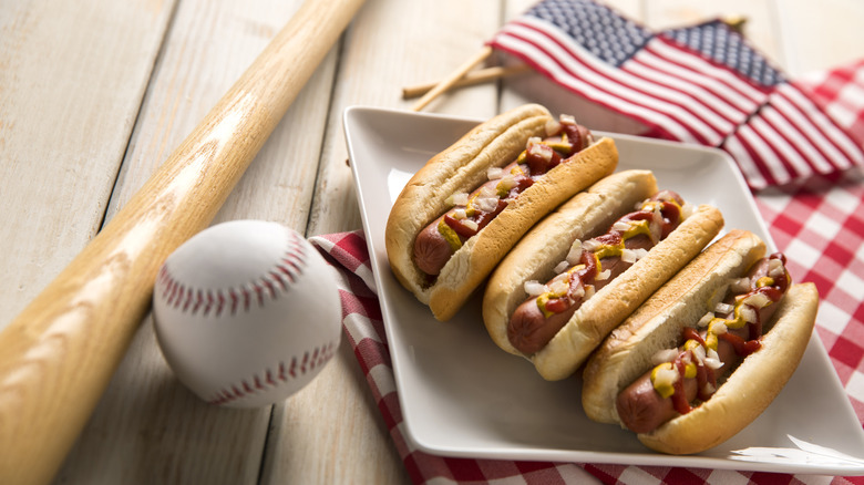 Three hot dogs on a plate topped with ketchup mustard and onion next to a baseball and a bat