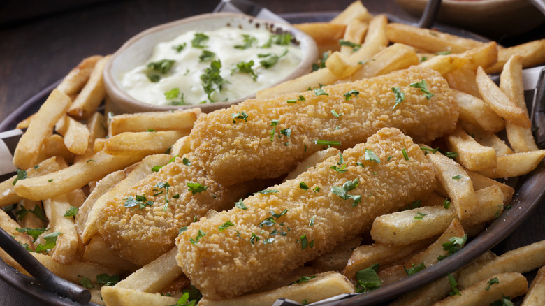 A close-up of fried fish pieces on top of chips, and including a bowl of tartar sauce