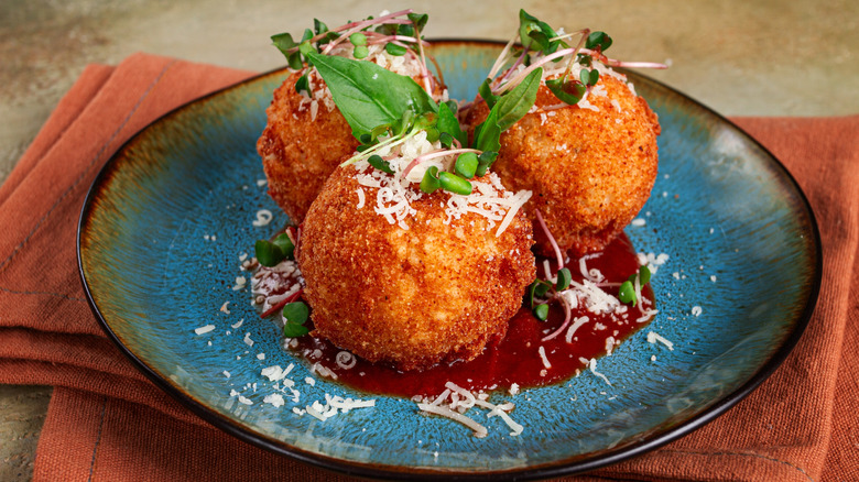 Three deep-fried arancini sit atop some tomato sauce on a plate, dressed with parmesan and greens