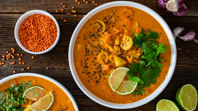 Bowls of red lentil soup topped with cilantro and lime sit on a wooden table next to a bowl of uncooked red lentils.