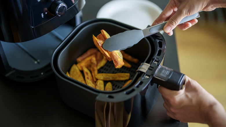 Person taking crisp potato wedges from an air fryer basked with tongs
