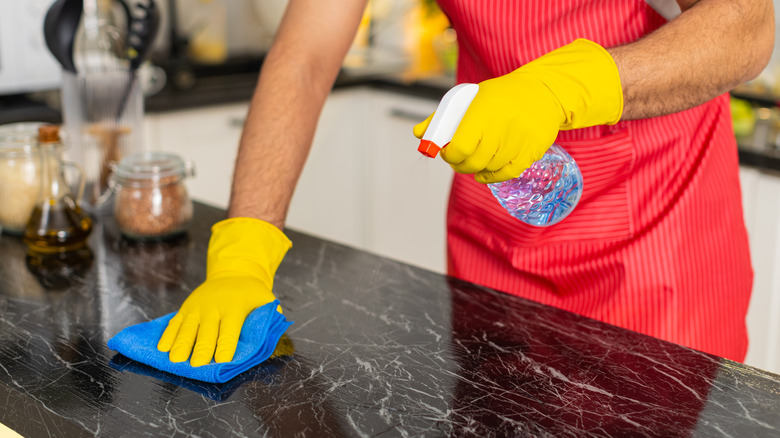 A person wearing yellow gloves sprays cleaner onto a black countertop and wipes it with a blue microfiber cloth.