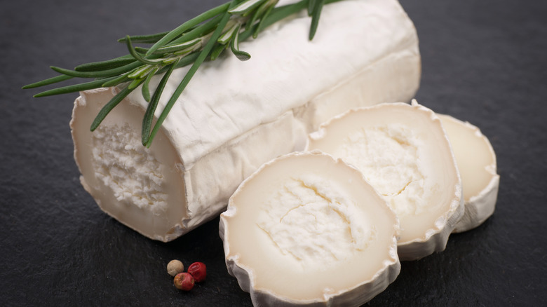 Slices next to log of aged goat cheese, topped with fresh herbs, lying on a grey surface