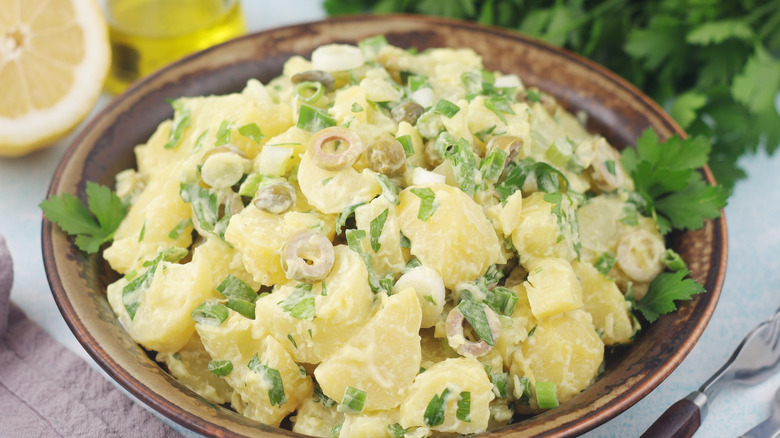 A bowl of creamy potato salad, with a sliced lemon and some fresh herbs in the background