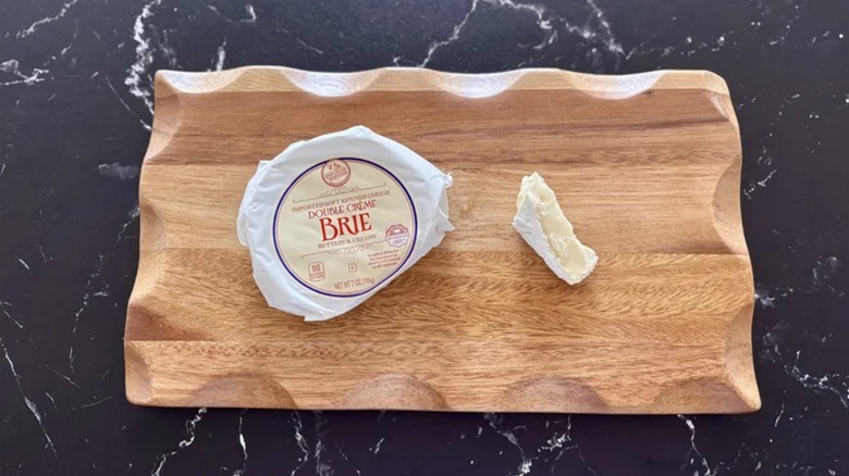 a wheel of Aldi brie in the wrapper next to a chunk of Brie on a wooden board