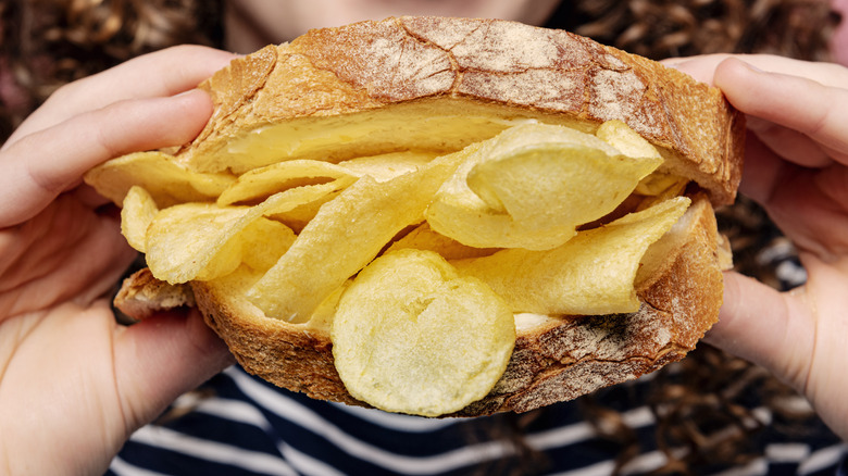 Hands holding a sandwich made with crusty looking bread that is bulging with potato chips