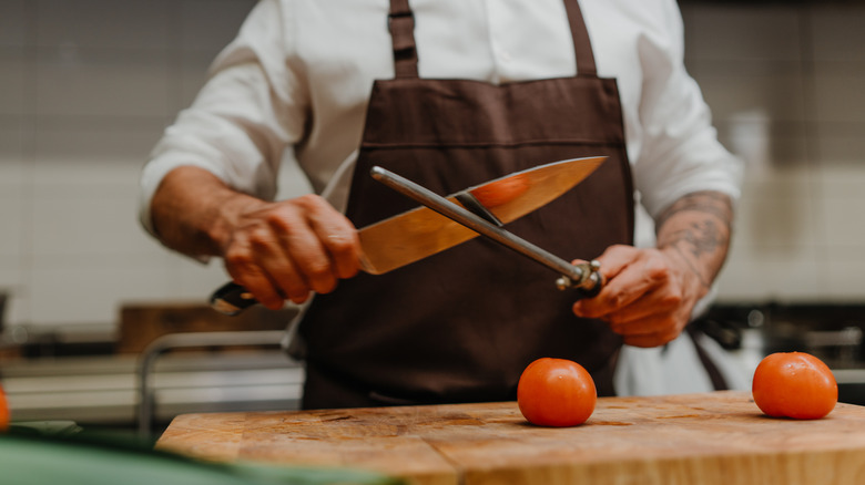 Person in an apron honing a chef's knife above a cutting board with tomatoes on it