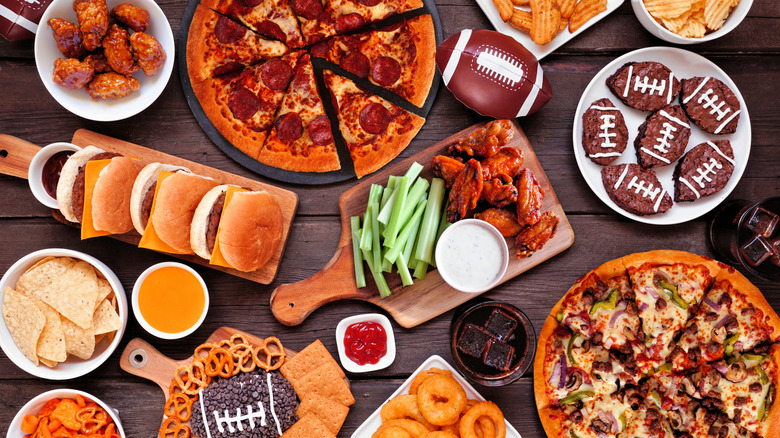 A table full of game night snacks, such as burgers, wings, pizza, football-shaped cupcakes, chips, soda and more.