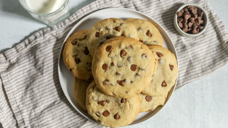 Stack of chocolate chip cookies on plate next to bowl of chocolate chips and glass of milk