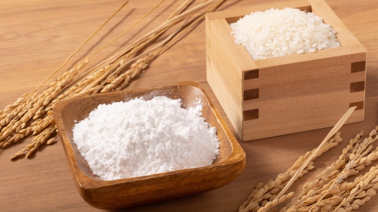 A wooden bowl of rice flour adjacent to another wooden container of rice, sitting on a wooden table with some dried grains