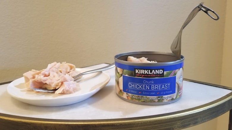 an open can of Kirkland chunk chicken breast  next to some of the meat on a white plate with a fork