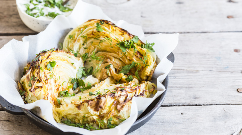 a paper-lined baking dish of roasted cabbage wedges topped with chopped fresh herbs on a wooden table with a bowl of sauce in the background