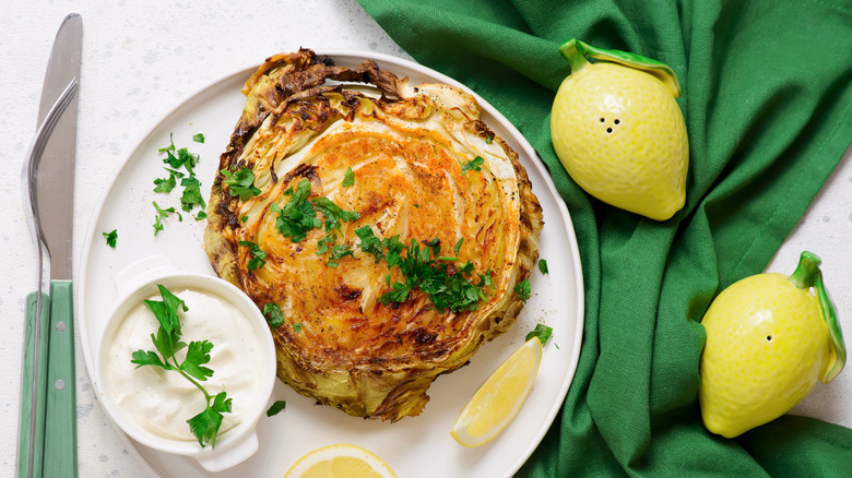 a roasted cabbage steak garnished with fresh herbs on a plate with a creamy sauce and lemon wedges