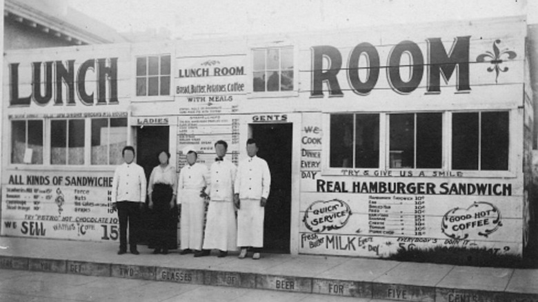 A black and white photo of a diner called Lunch Room with workers standing outside and the menu painted on the front of the building