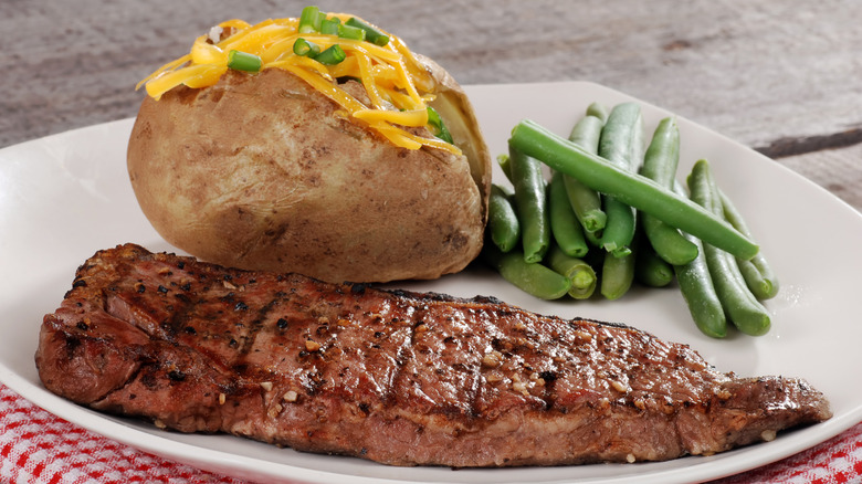 Grilled beef steak on white plate next to green beans and baked potato with cheese and scallions