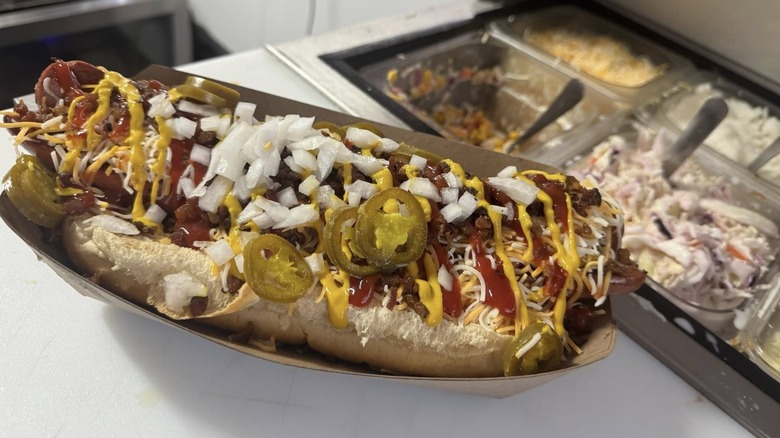 Giant hot dog loaded with ketchup, mustard, jalapenos, onions, and cheese in front of toppings