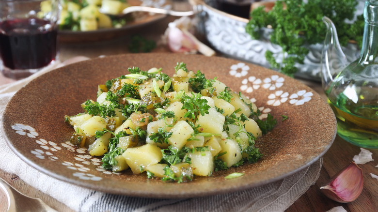 A rustic shallow bowl of potato salad with parsley and oil and wine on the side.