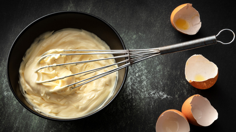 A bowl of homemade mayonnaise with a whisk in it and broken egg shells beside it