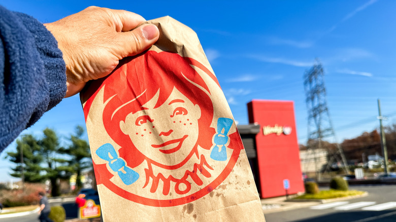 Hand holding up a Wendy's takeout bag in front of a drive-thru