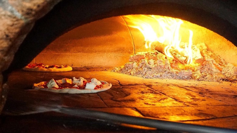 Two pizzas in a woodfired pizza oven with a pizza peel in the foreground
