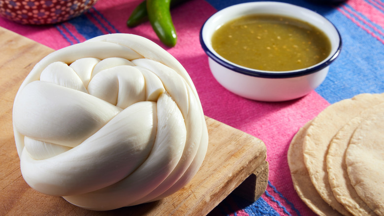 A large ball of queso de Oaxaca sits near a bowl of green salsa and a stack of tortillas