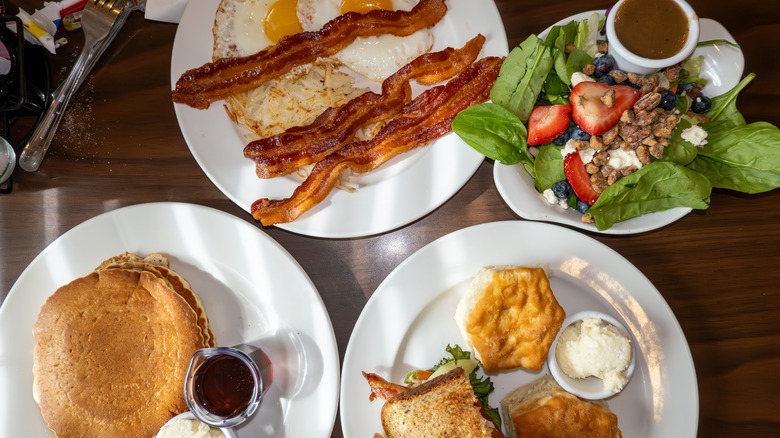 Four plates of diner food; eggs, bacon, pancakes, strawberries, biscuit, toast
