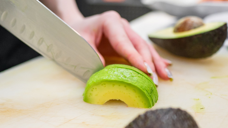 Hand actively cutting halved avocado in front of the other sliced half on a cutting board.